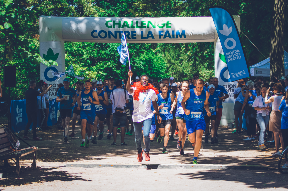 Des participants prennent le départ d’une course solidaire sous une arche gonflable portant l’inscription 'Challenge contre la faim'. Un homme souriant en chemise blanche et boa rouge court en tête en brandissant un drapeau, suivi par un groupe de coureurs en tenue de sport bleue. L’événement se déroule en extérieur, dans un parc boisé, devant un public rassemblé.