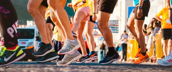 Gros plan sur les pieds de plusieurs coureurs en pleine course sur route, avec des baskets colorées, lors d’une course solidaire en extérieur sous le soleil, entourés de spectateurs.